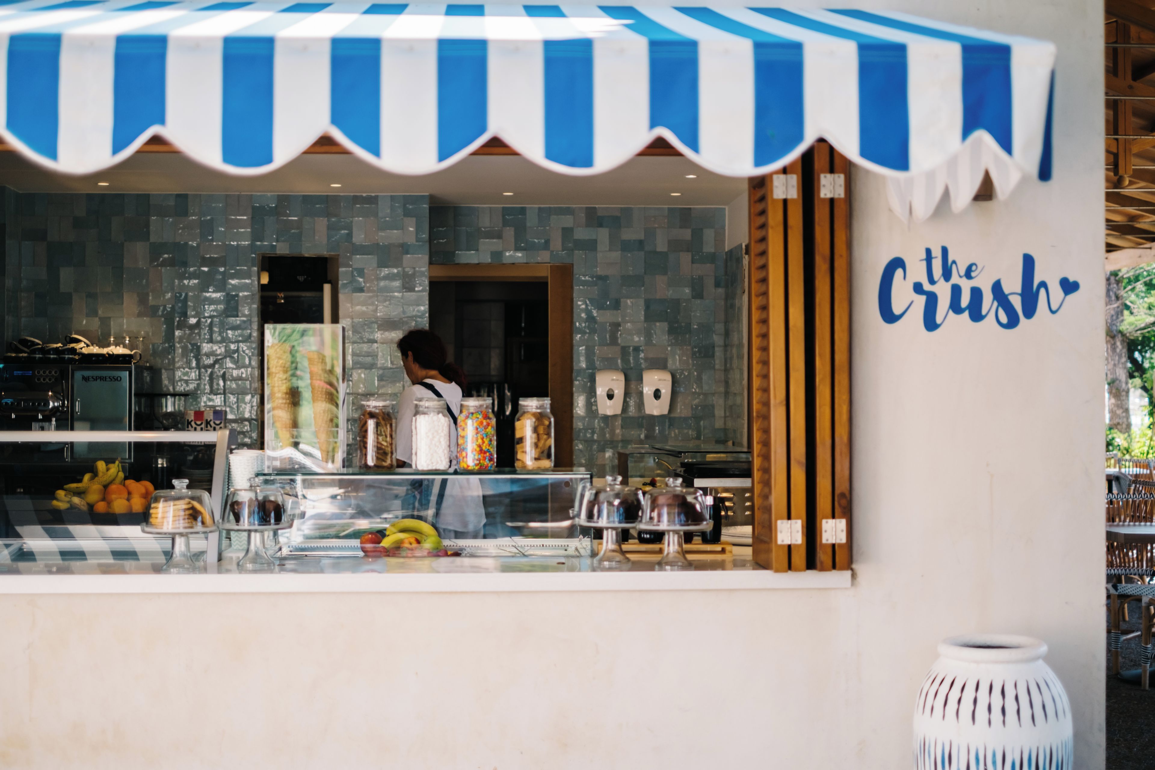 Una cafetería con toldo a rayas, un mostrador con diferentes productos alimenticios y una pared con un letrero.