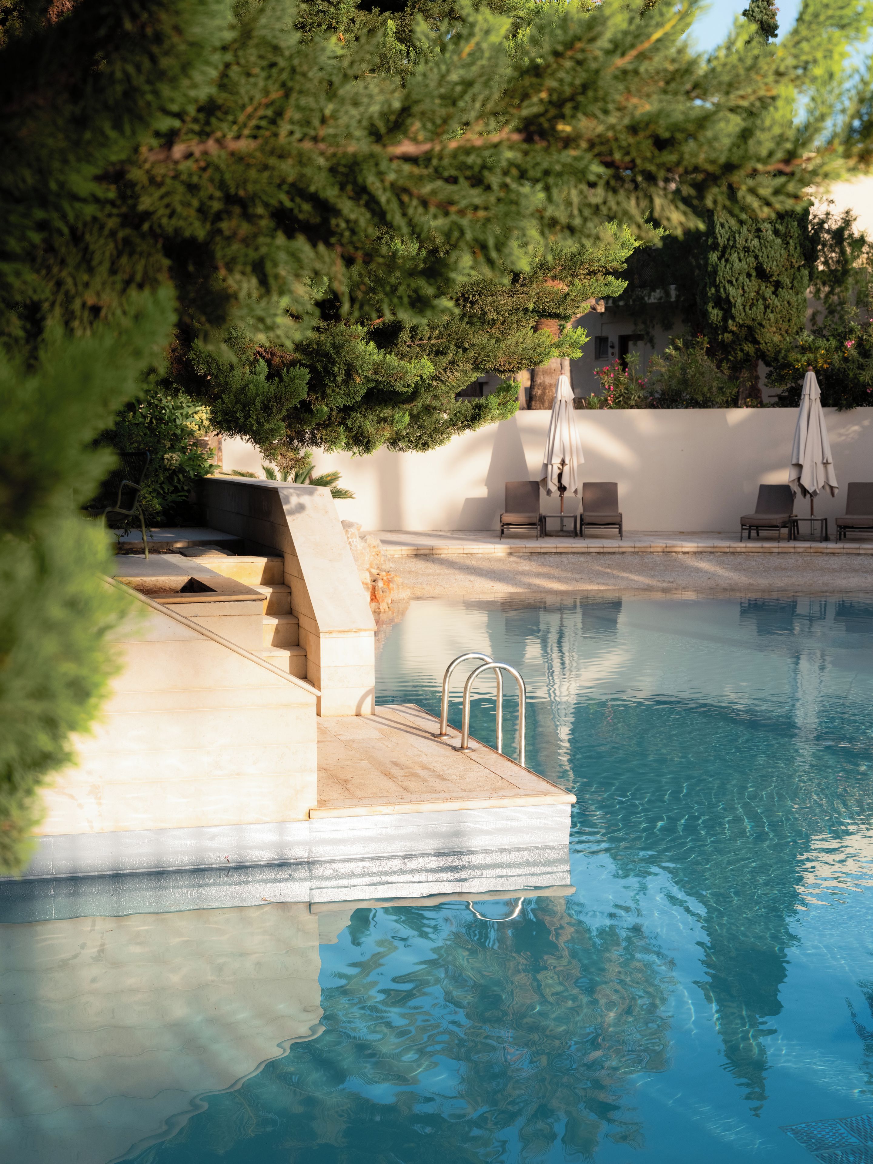 Una piscina amb superfície d'aigua cristal·lina, un conjunt d'escales que hi baixen i diverses gandules i ombrel·les al voltant de la zona de la piscina.