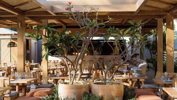 Un área de comedor al aire libre con mesas y sillas de madera, rodeada de plantas en macetas y una pérgola de madera.