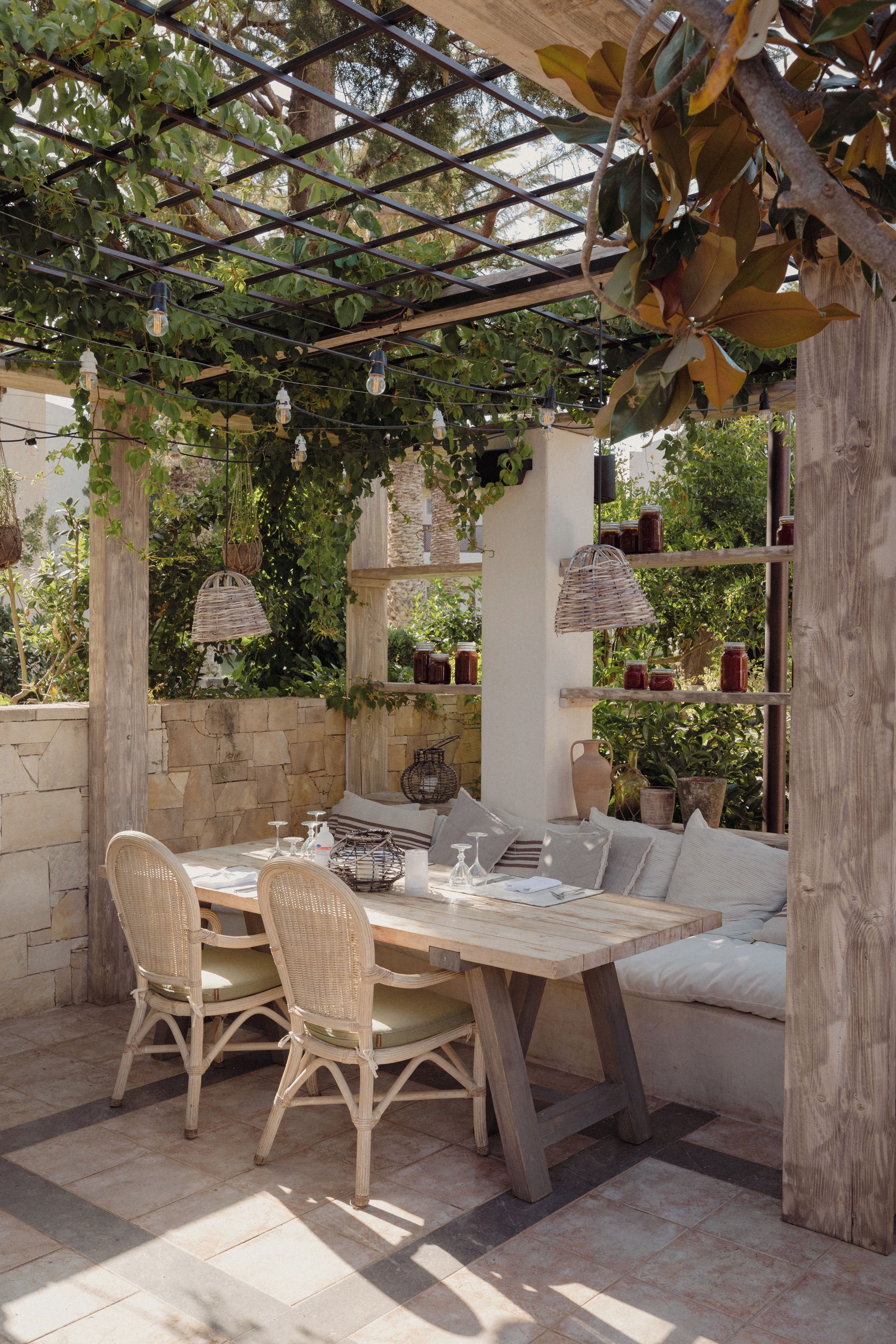 An outdoor patio with a wooden table and wicker chairs, surrounded by greenery and stone walls.