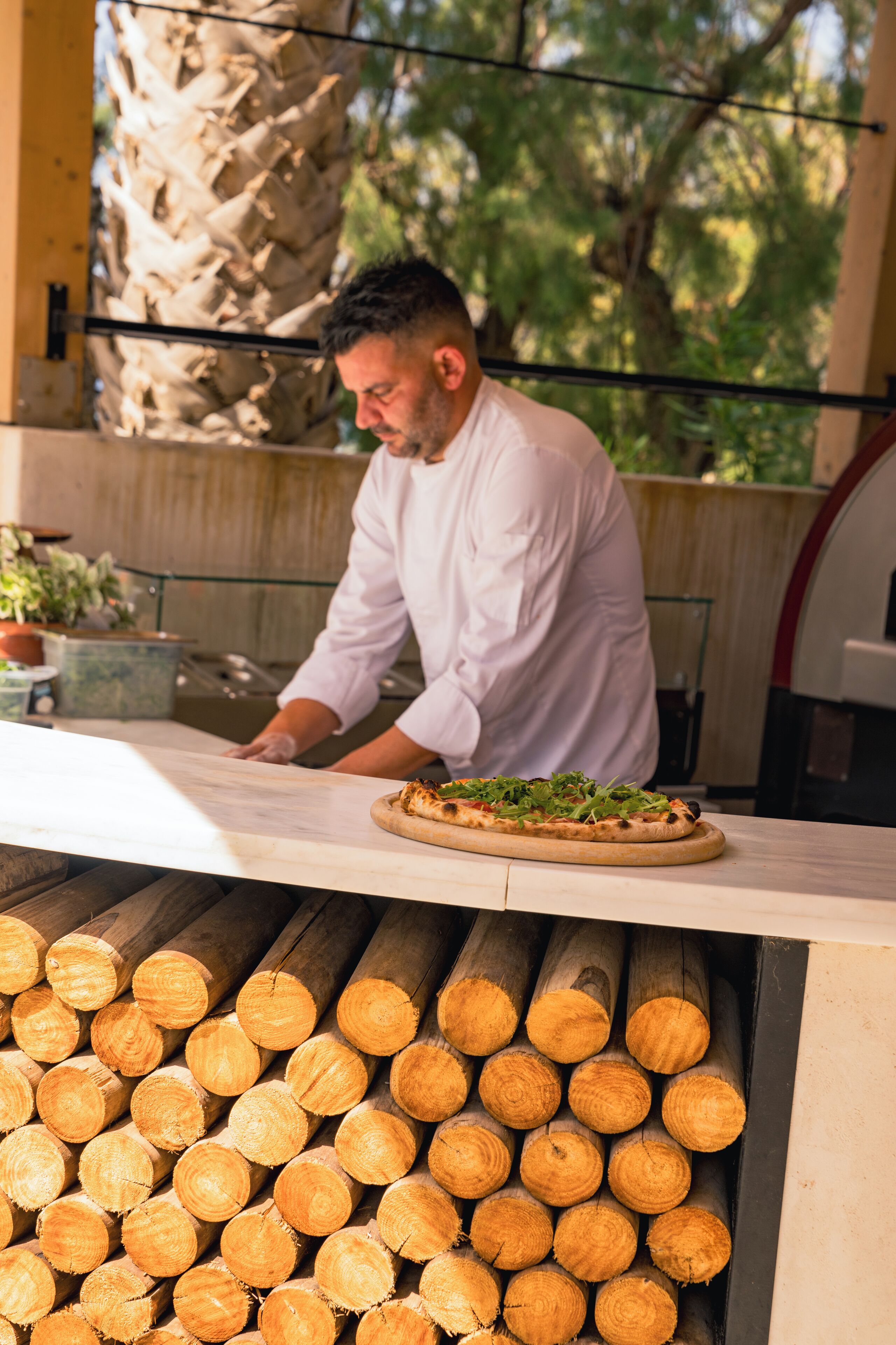 Un cocinero preparando comida en una cocina, con una pizza sobre la encimera y una pila de leña al fondo.