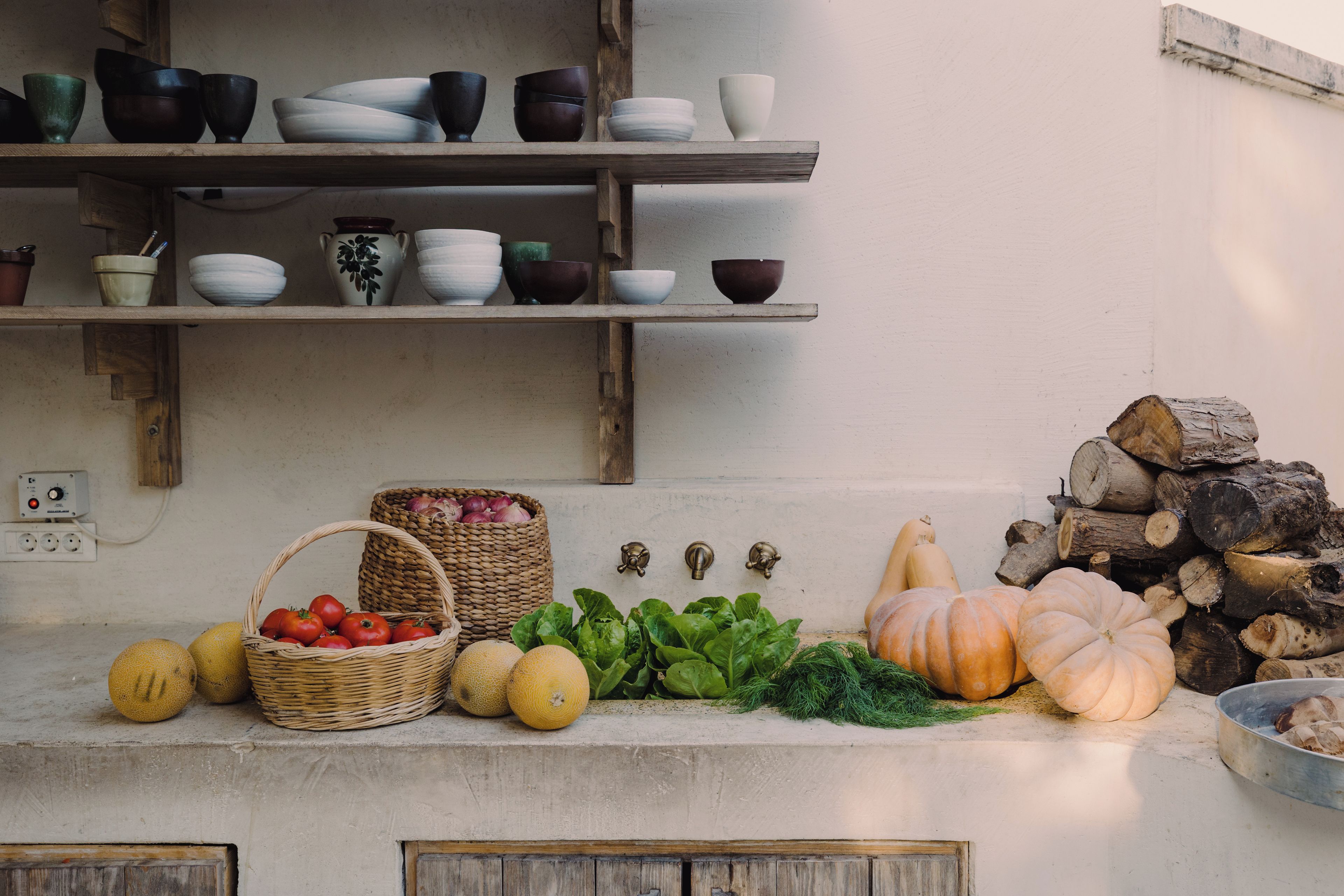 A kitchen counter with various bowls, a basket of tomatoes, and a pile of firewood.
