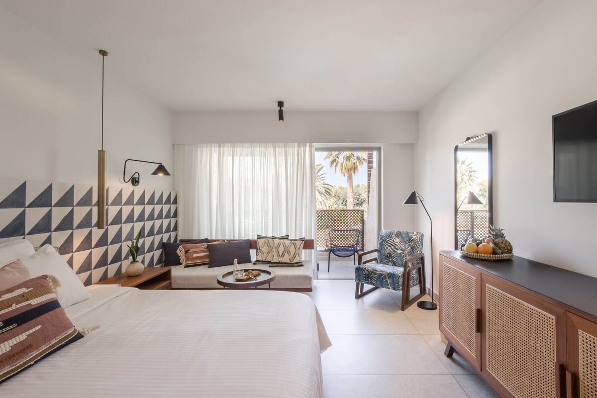 Ein modernes Hotelzimmer mit einem großen Bett, einer Couch, einer Essecke und einem Balkon mit Blick auf Palmen.
