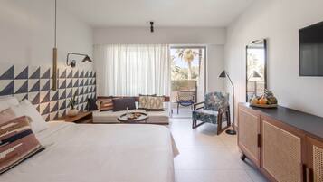 A modern hotel room with a large bed, a sofa, a dining area, and a balcony with a view of palm trees.