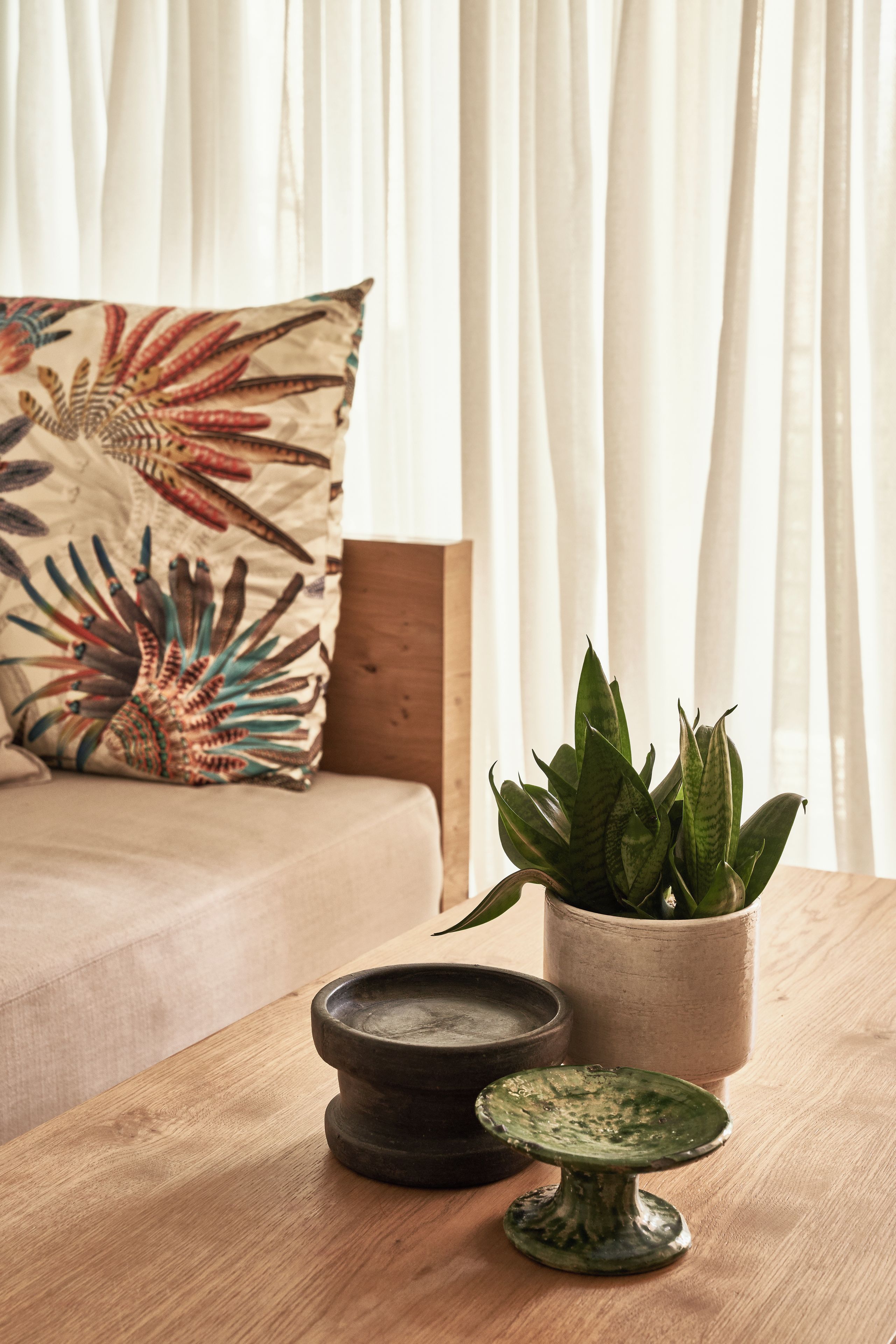 A room with a bed, a pillow with a leaf pattern, a potted plant, and a small table with a bowl and a decorative item.