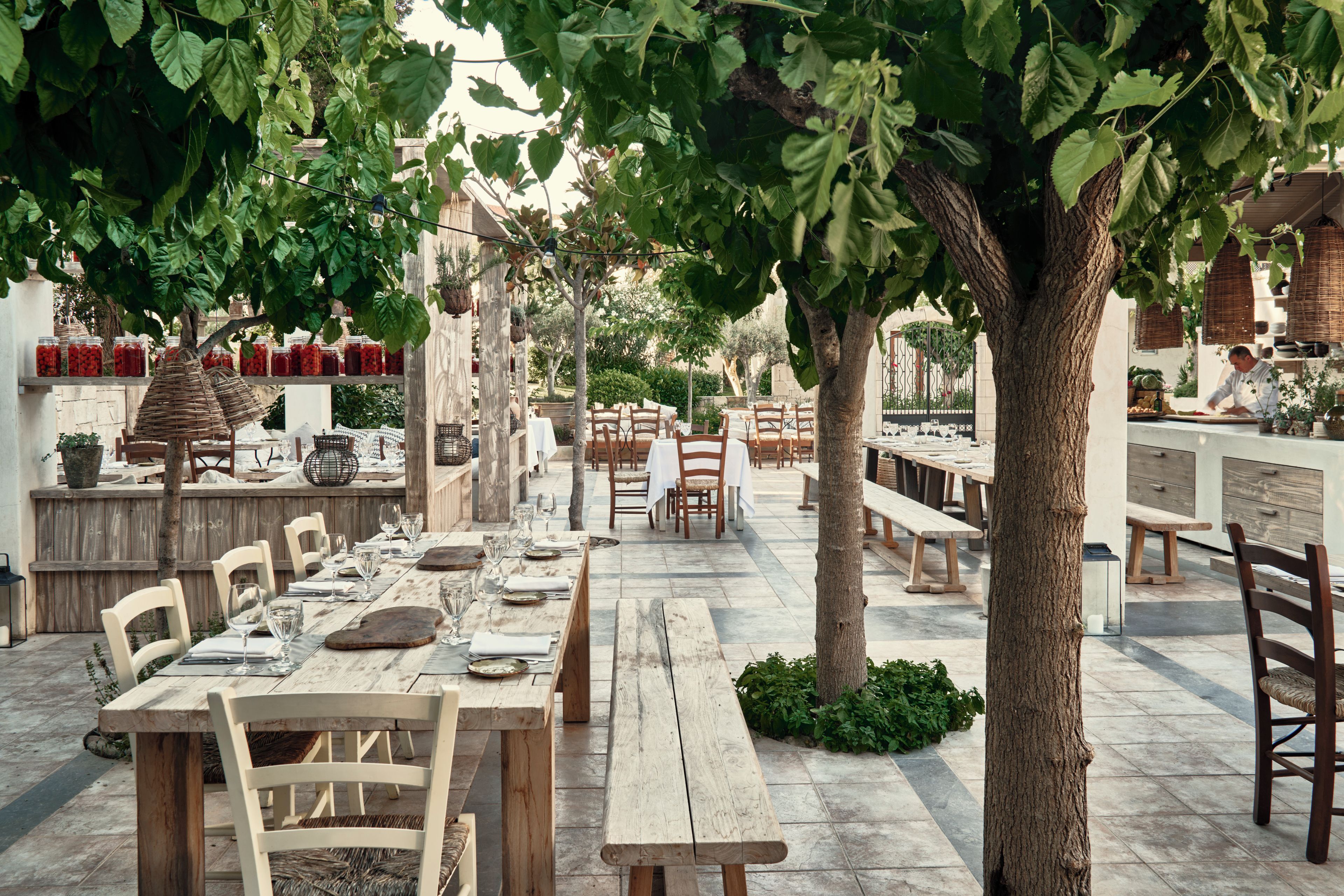 Un área de comedor al aire libre con mesas y sillas de madera, rodeada de vegetación y suelo de piedra.