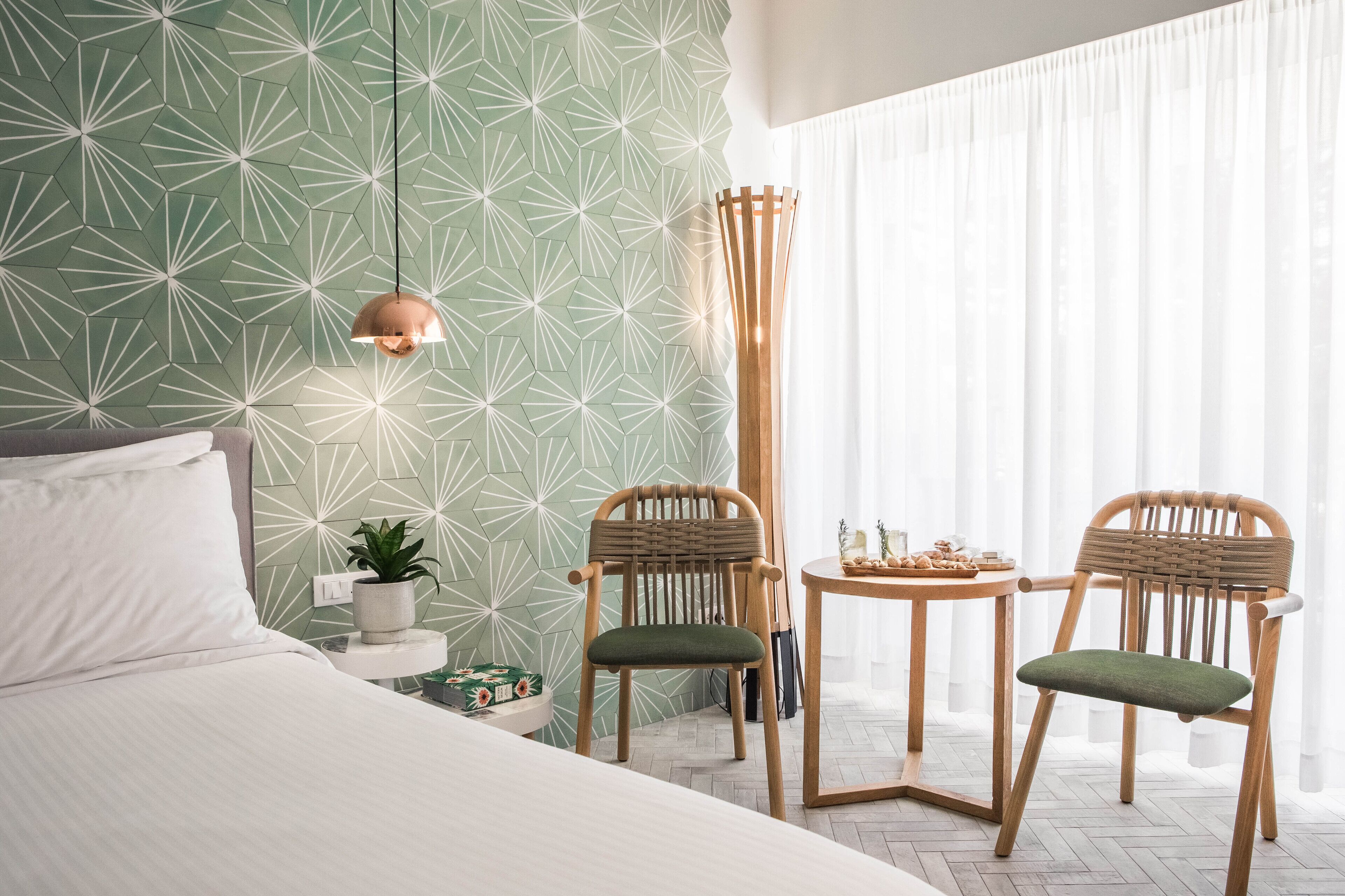 Une chambre d’hôtel moderne avec un mur orné d’un motif géométrique, un lit, deux chaises, une petite table avec des en-cas et un grand lampadaire.