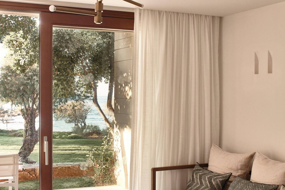 Ein modernes Wohnzimmer mit einer Couch, einem Glastisch und Blick auf einen Garten und Bäume durch eine Glasschiebetür.
