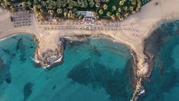 Vue aérienne d’un complexe hôtelier en bord de mer, avec plusieurs rangées de parasols et de chaises longues, un parking et un bâtiment doté d’un toit.