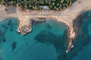 Vue aérienne d’un complexe hôtelier en bord de mer, avec plusieurs rangées de parasols et de chaises longues, un parking et un bâtiment doté d’un toit.