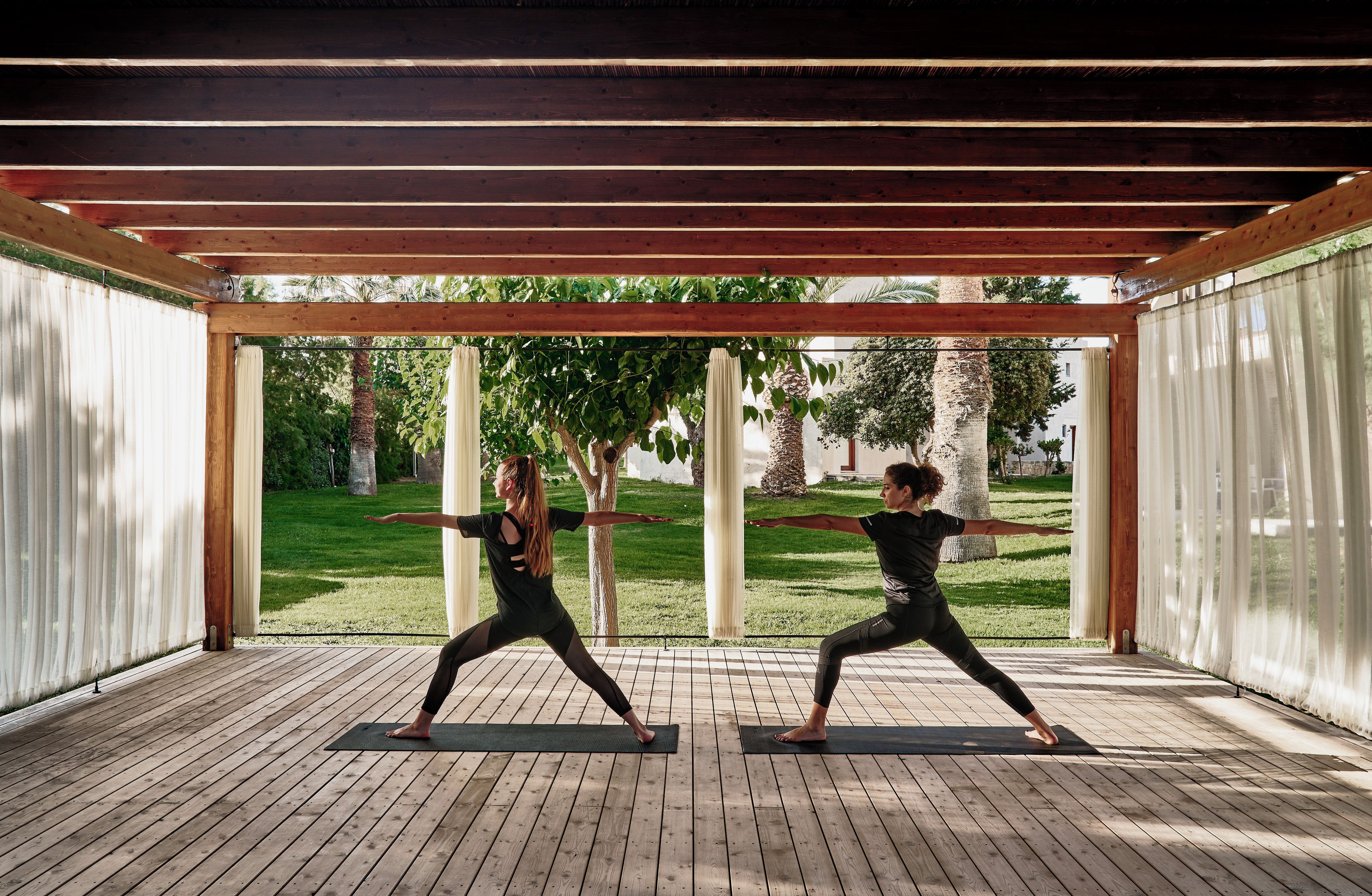 Dos personas practicando yoga en un espacio al aire libre cubierto, con piso de madera y vista a un paisaje con césped.