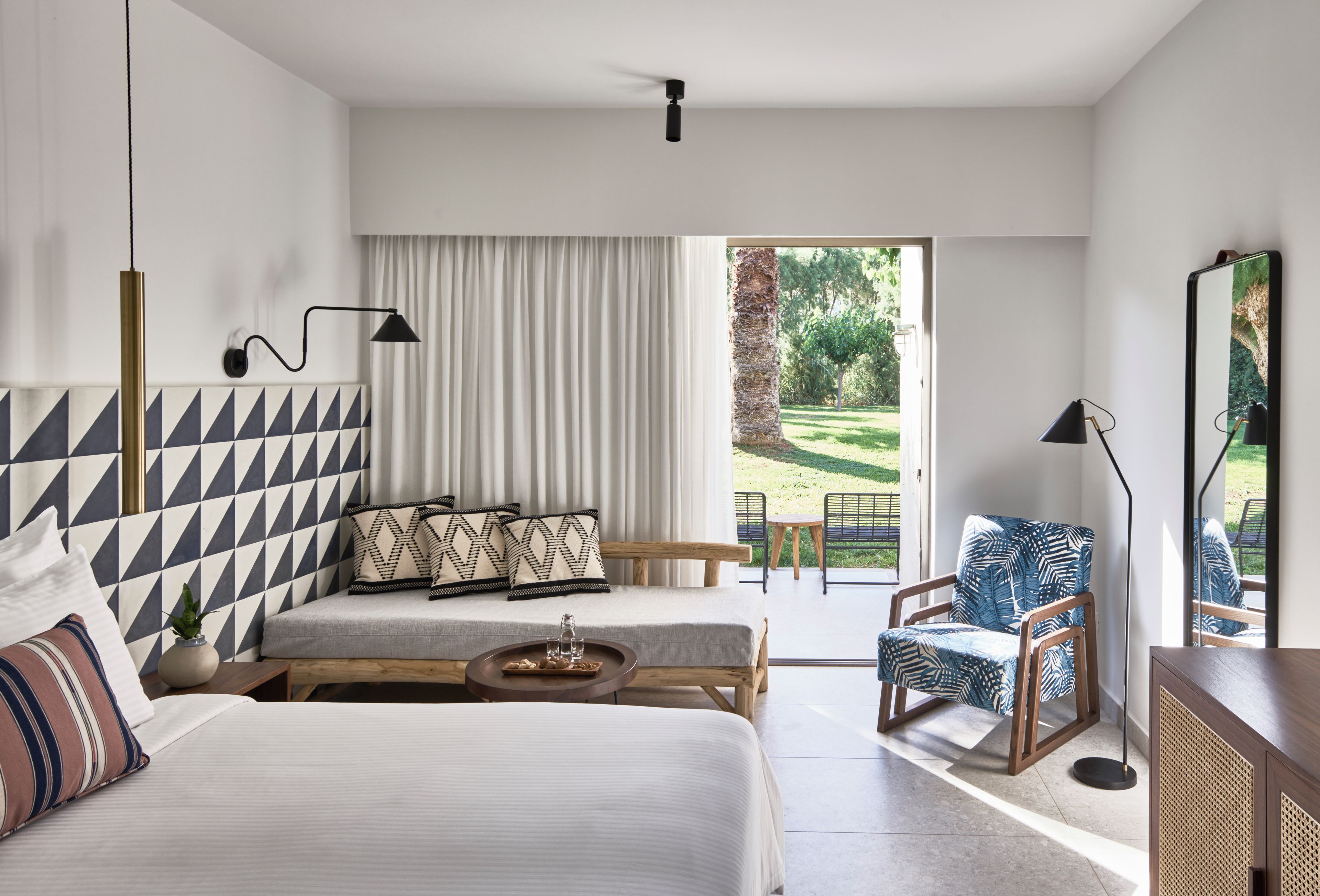 Une chambre d’hôtel moderne dotée d’un grand lit, d’un canapé, d’un fauteuil, d’une petite table et offrant une vue sur un jardin grâce à une porte coulissante.