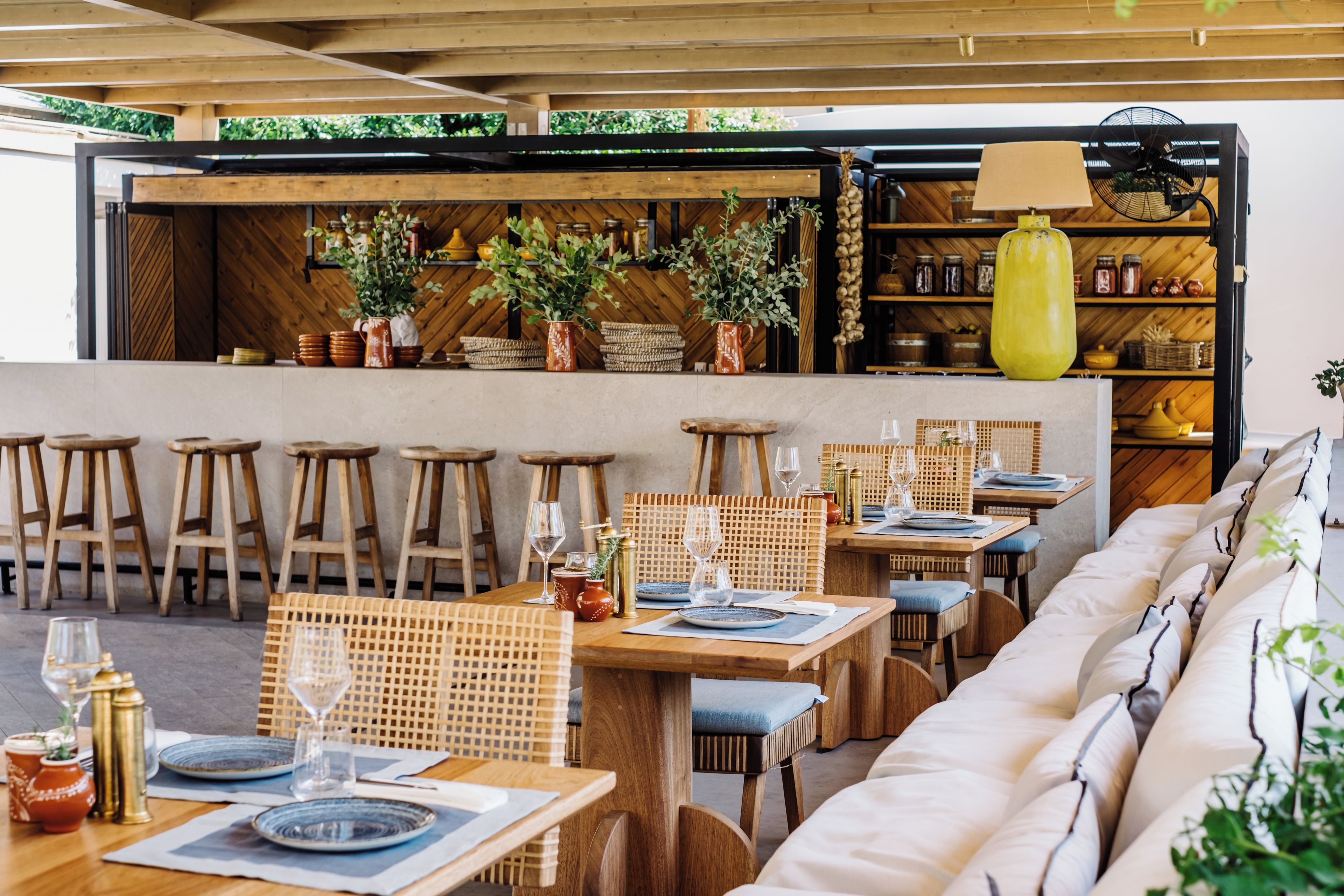 Un restaurante con mesas de madera, taburetes de bar y una zona de bar con estanterías.