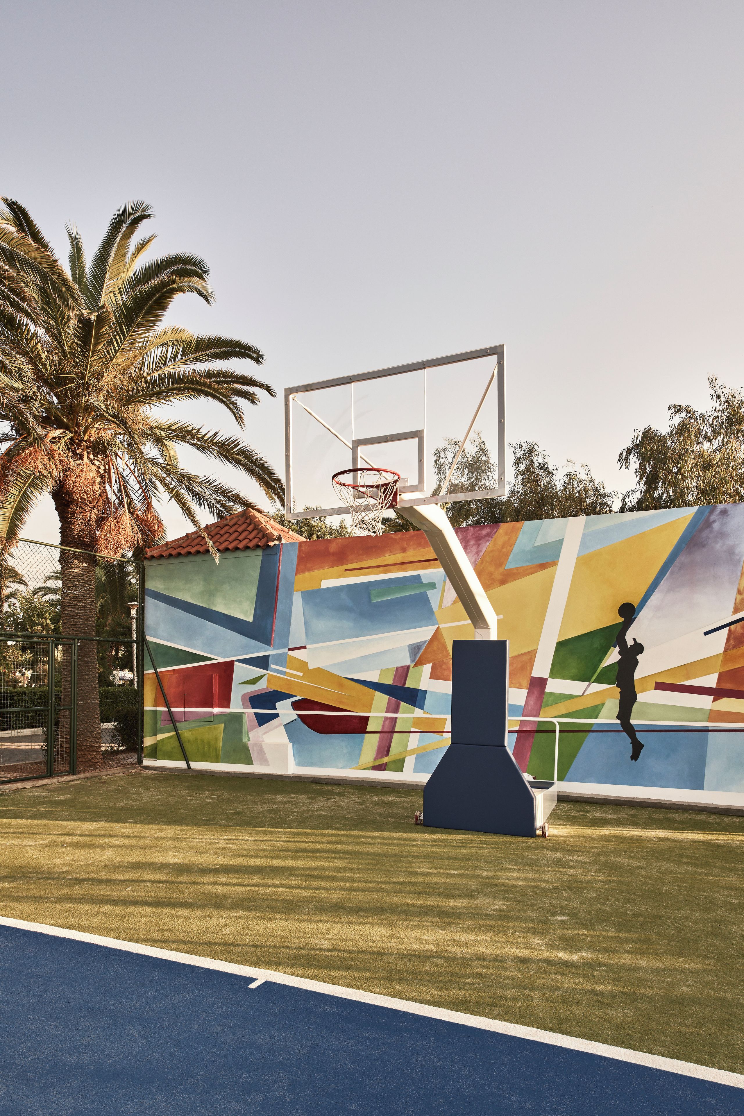 An outdoor basketball court with a colorful mural, a basketball hoop, and a palm tree.