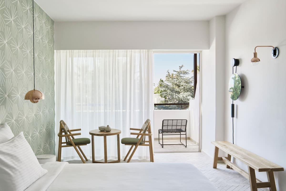 Ein Schlafzimmer mit einem Bett, zwei Stühlen, einem kleinen Tisch, einer Bank und einem Balkon mit Blick auf Bäume.