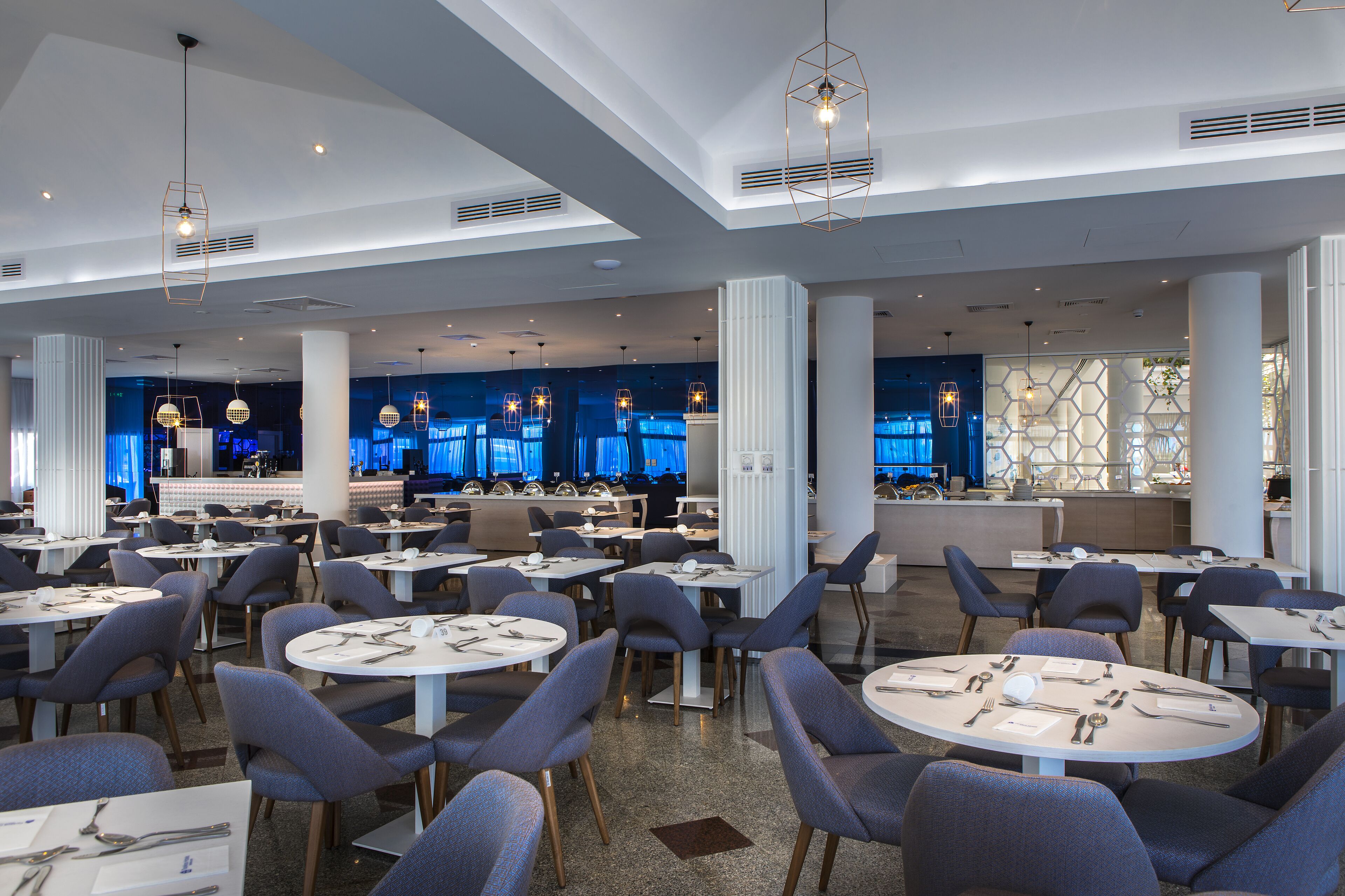 A spacious dining area with round tables and modern chairs, a central bar area, and a high ceiling with skylights.