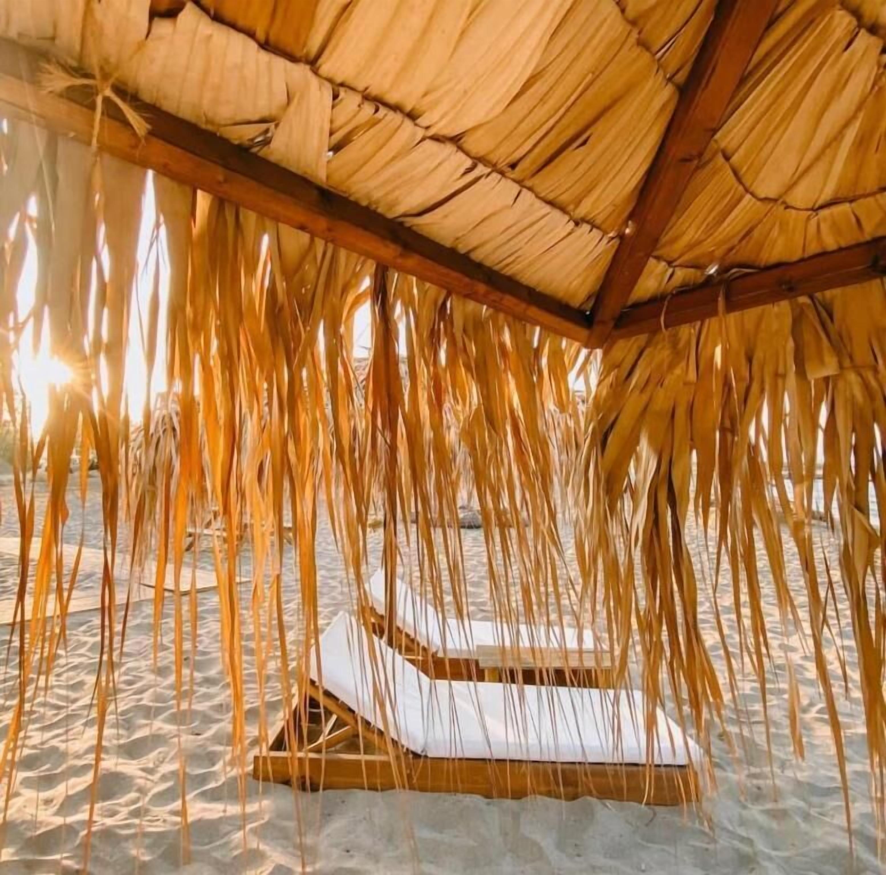 A thatched-roof structure with a wooden bench and a woven mat on the sand floor.