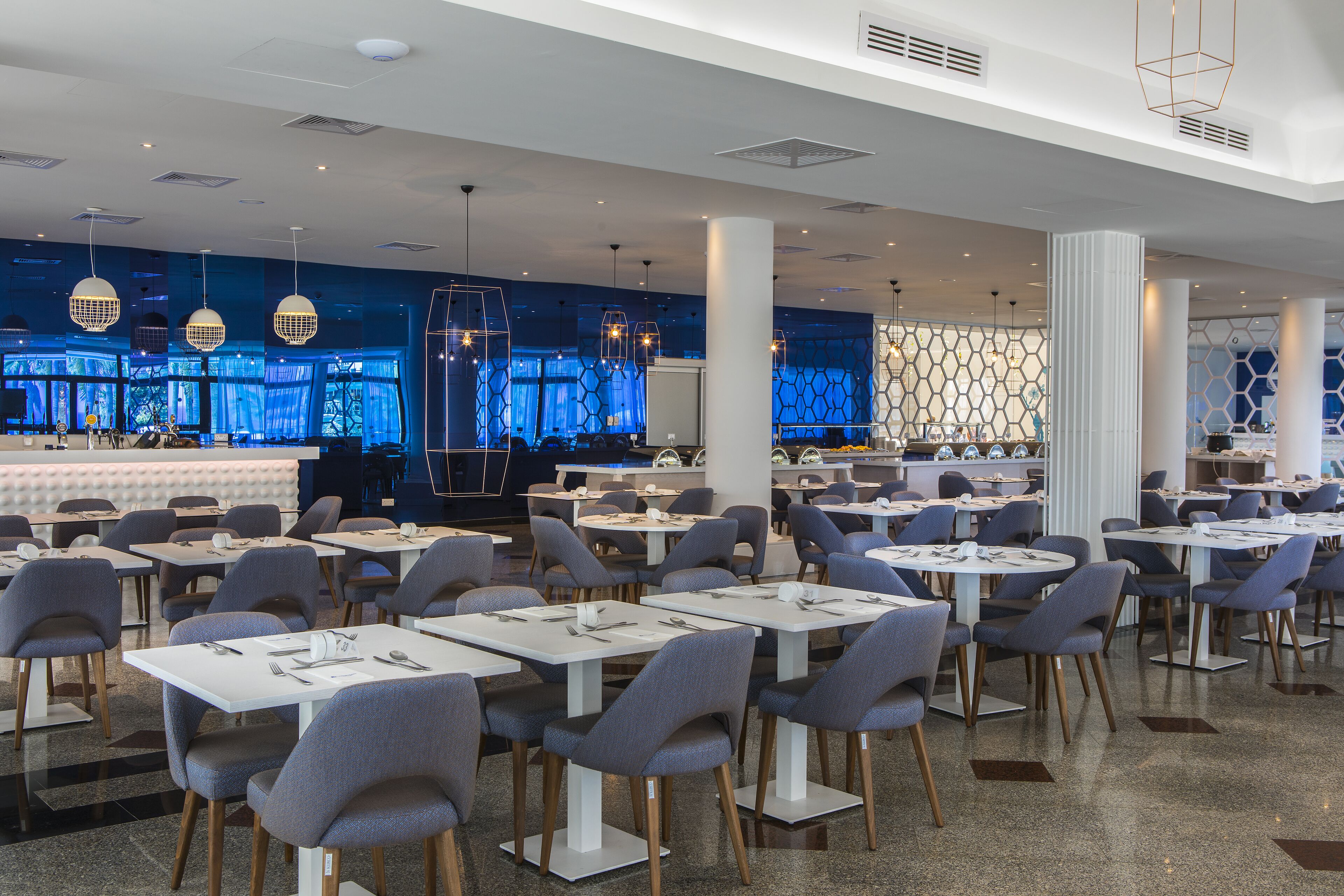 Un restaurante moderno con mesas redondas y sillas grises, una zona de bar con iluminación azul y una pared decorativa con patrones hexagonales.