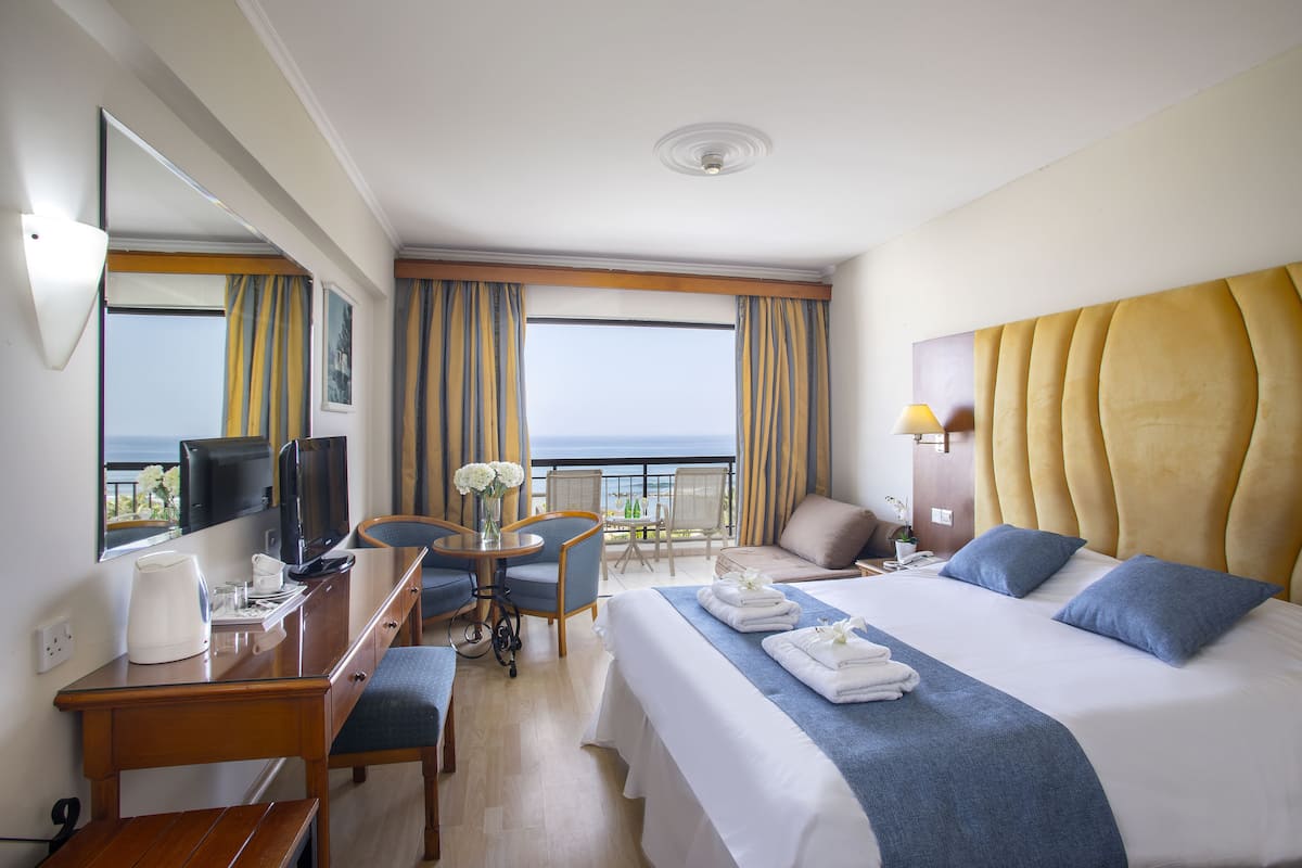 A hotel room with a large bed, a desk with a chair, a small table with chairs, a TV, and a balcony with a view of the sea.