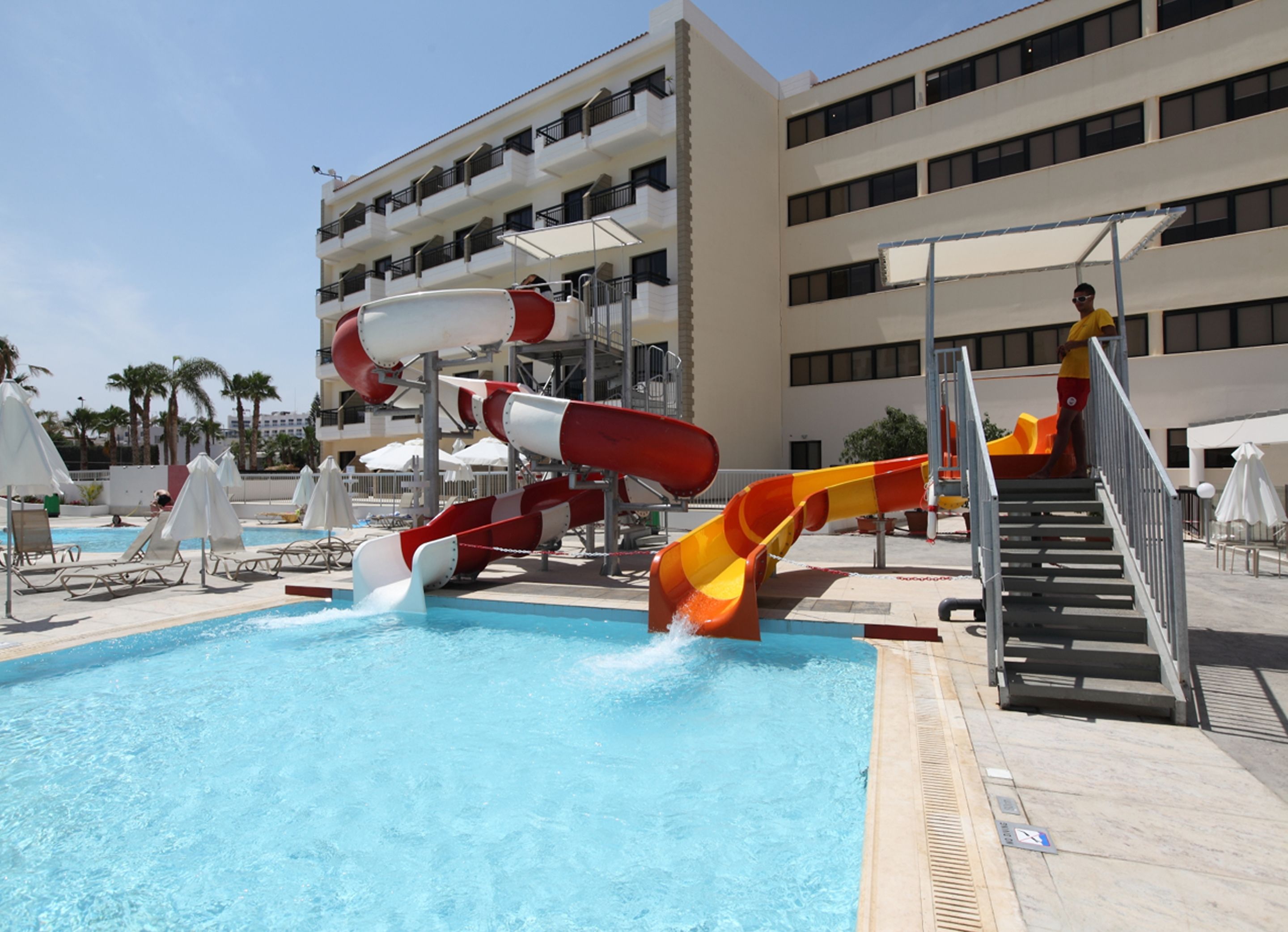 A swimming pool with multiple water slides, a staircase, and a building with balconies in the background.