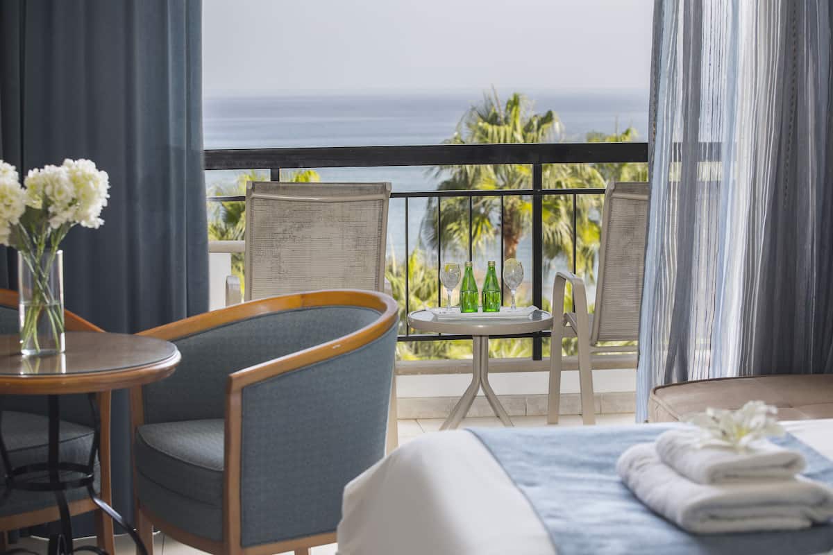 A hotel room with a balcony, a bed with a blue bedspread, a chair, a small table with a vase of flowers, and a view of the ocean.