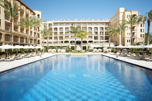 A large pool with lounge chairs and umbrellas, surrounded by a hotel with multiple floors and balconies.