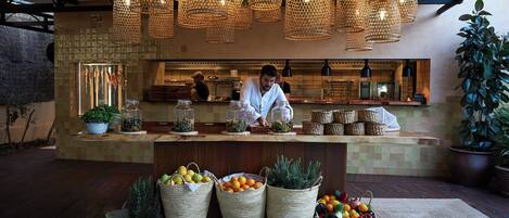 A restaurant with wicker baskets filled with fruits and vegetables, hanging wicker lamps, and a chef preparing food.