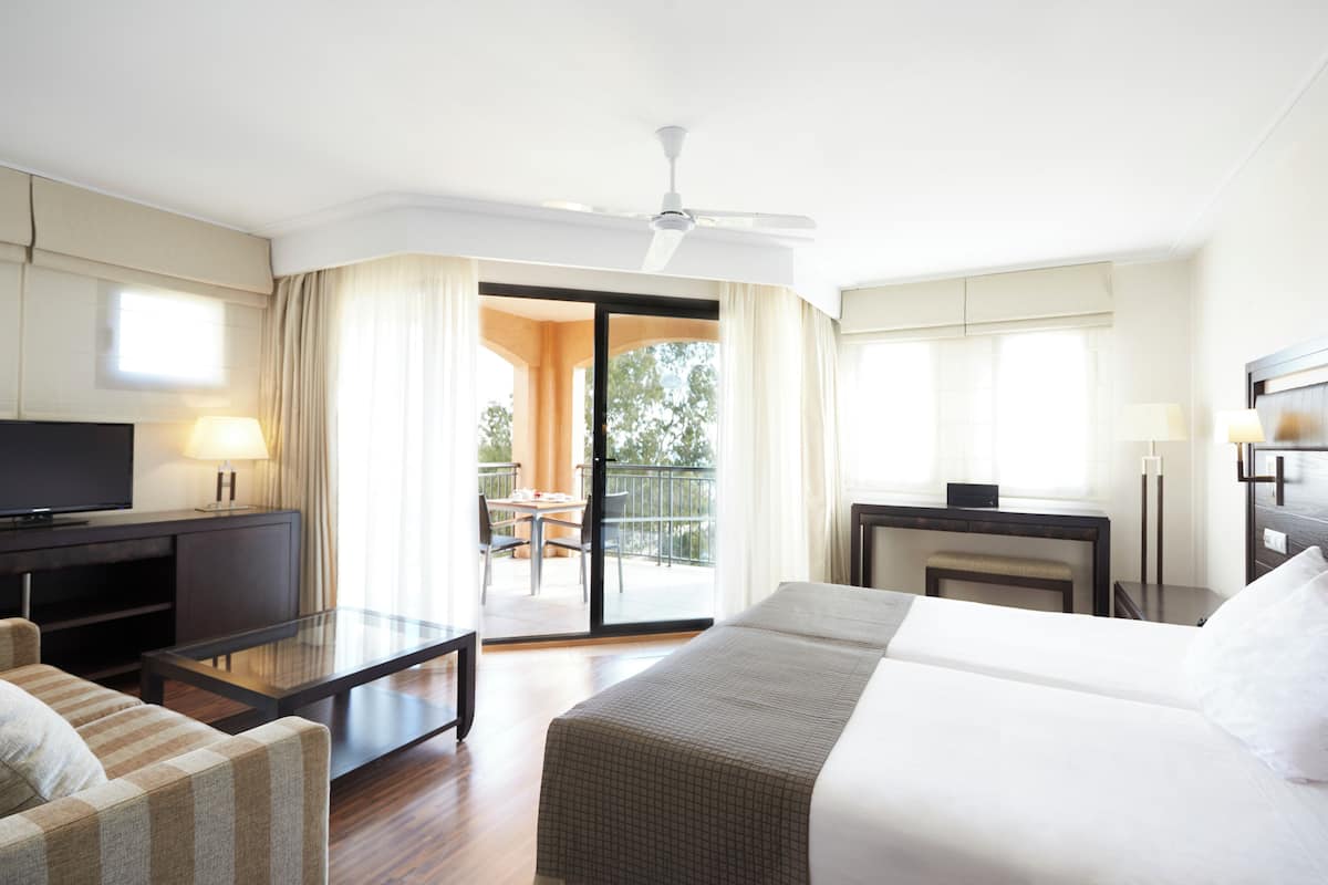 A hotel room with a bed, a TV, a glass table, a sofa, a desk, and a balcony with a table and chairs.