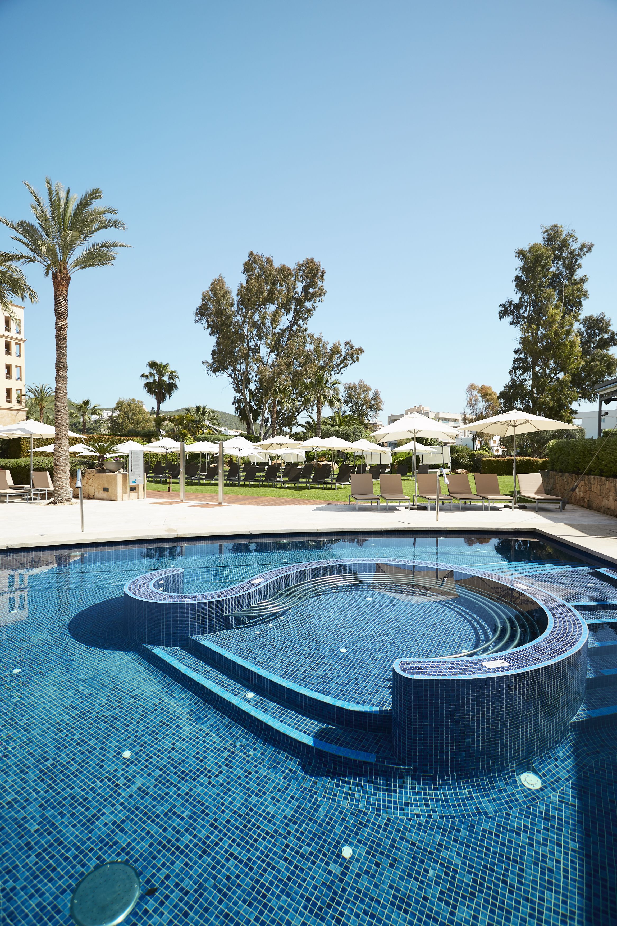 A swimming pool with a tiled design, surrounded by lounge chairs and umbrellas, with palm trees and buildings in the background.