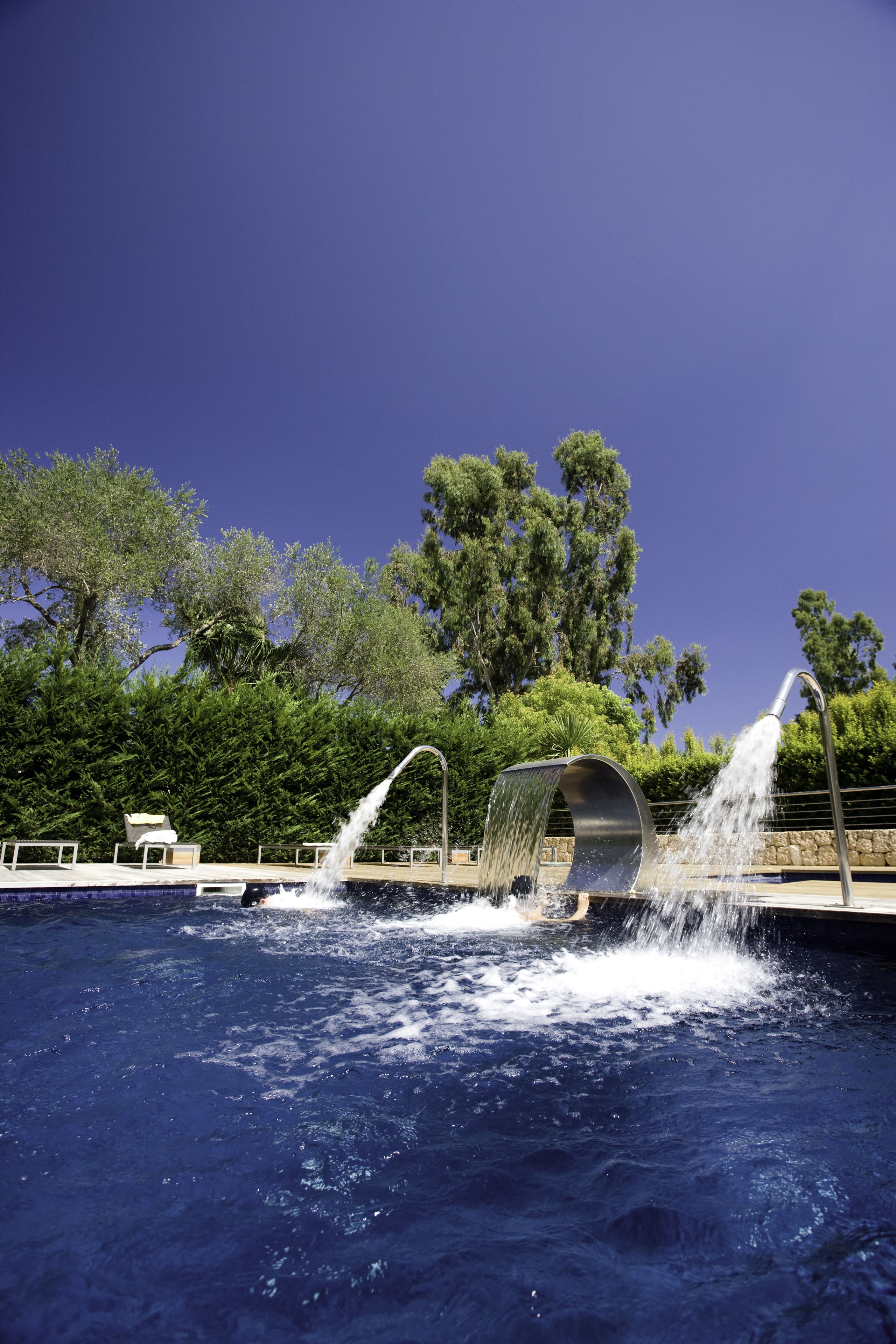 A swimming pool with a water feature, surrounded by trees and a clear sky.