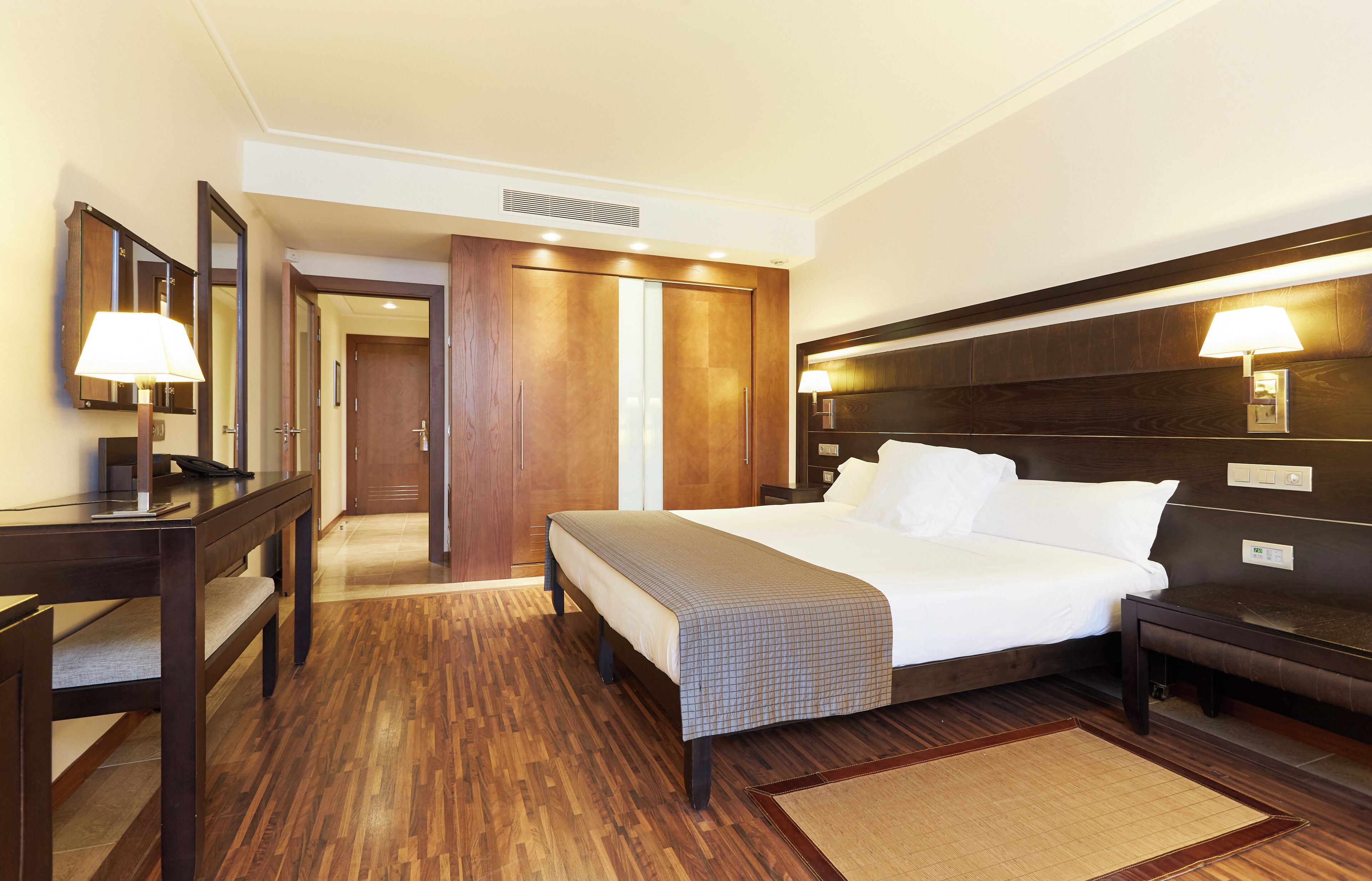 Quarto de hotel com cama, mesas de cabeceira, escrivaninha, espelho, guarda-roupa e abajur.