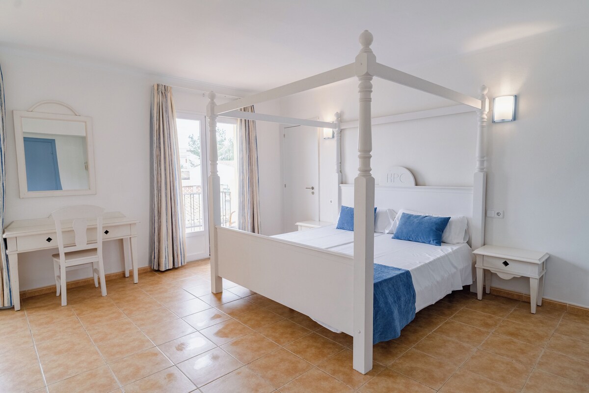 Un dormitorio con cama con dosel, un escritorio con sillas, una mesita de noche y un espejo.