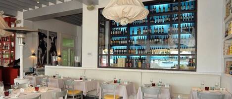 Interior de restaurante con mesas dispuestas para comer, una estantería con vino al fondo y un gran ventanal con vista a la calle.