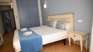 A neatly made bed with a blue bedspread, a wooden nightstand with a drawer, and a blue wall with a light switch.
