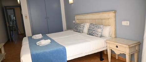 A neatly made bed with a blue bedspread, a wooden nightstand with a drawer, and a blue wall with a light switch.
