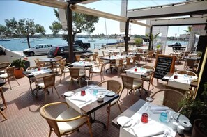 Un área de comedor al aire libre con mesas y sillas, con vistas a un puerto deportivo con barcos.