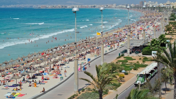 Una playa con sombrillas y gente, una carretera con un autobús, y edificios al fondo.