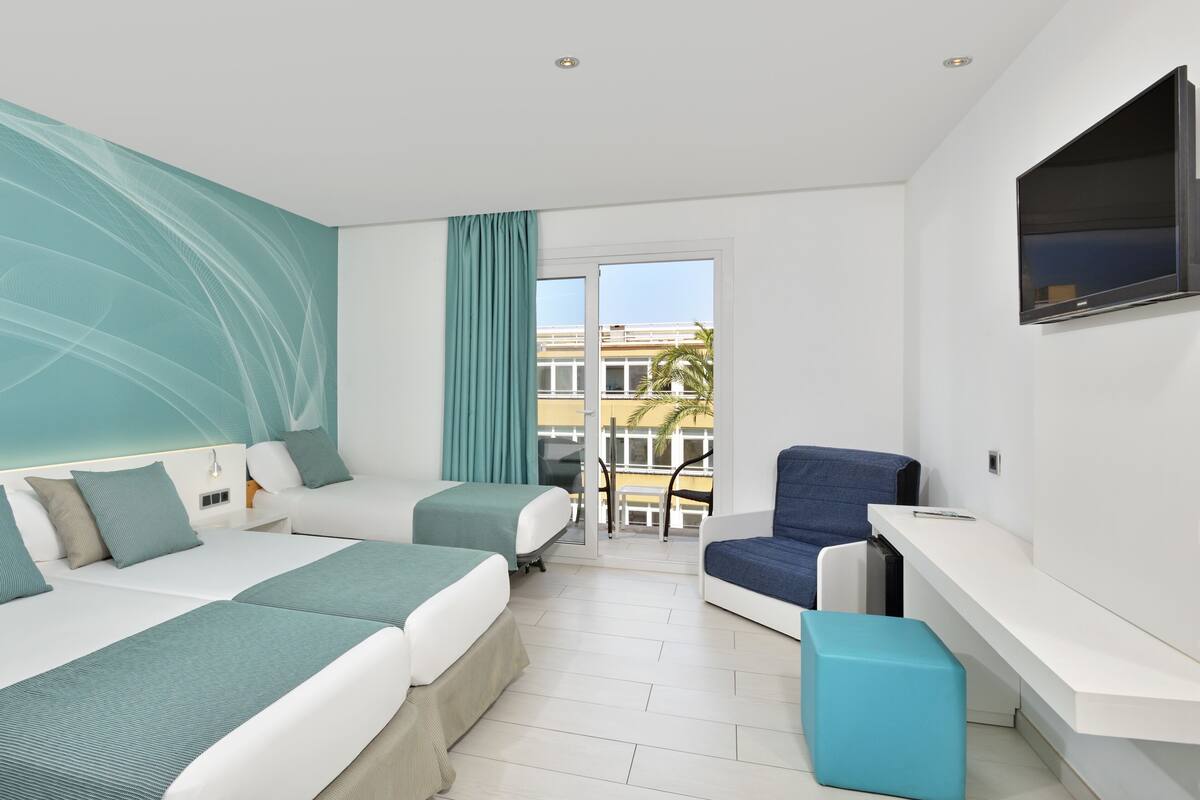 Ein modernes Hotelzimmer mit einem großen Bett, einem Schreibtisch, einem Sessel und einem Balkon mit Blick auf Gebäude und Palmen.