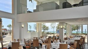 Un restaurante con varias mesas dispuestas para comer, ventanas amplias que ofrecen vistas a la playa y al océano, y un diseño de interiores moderno.