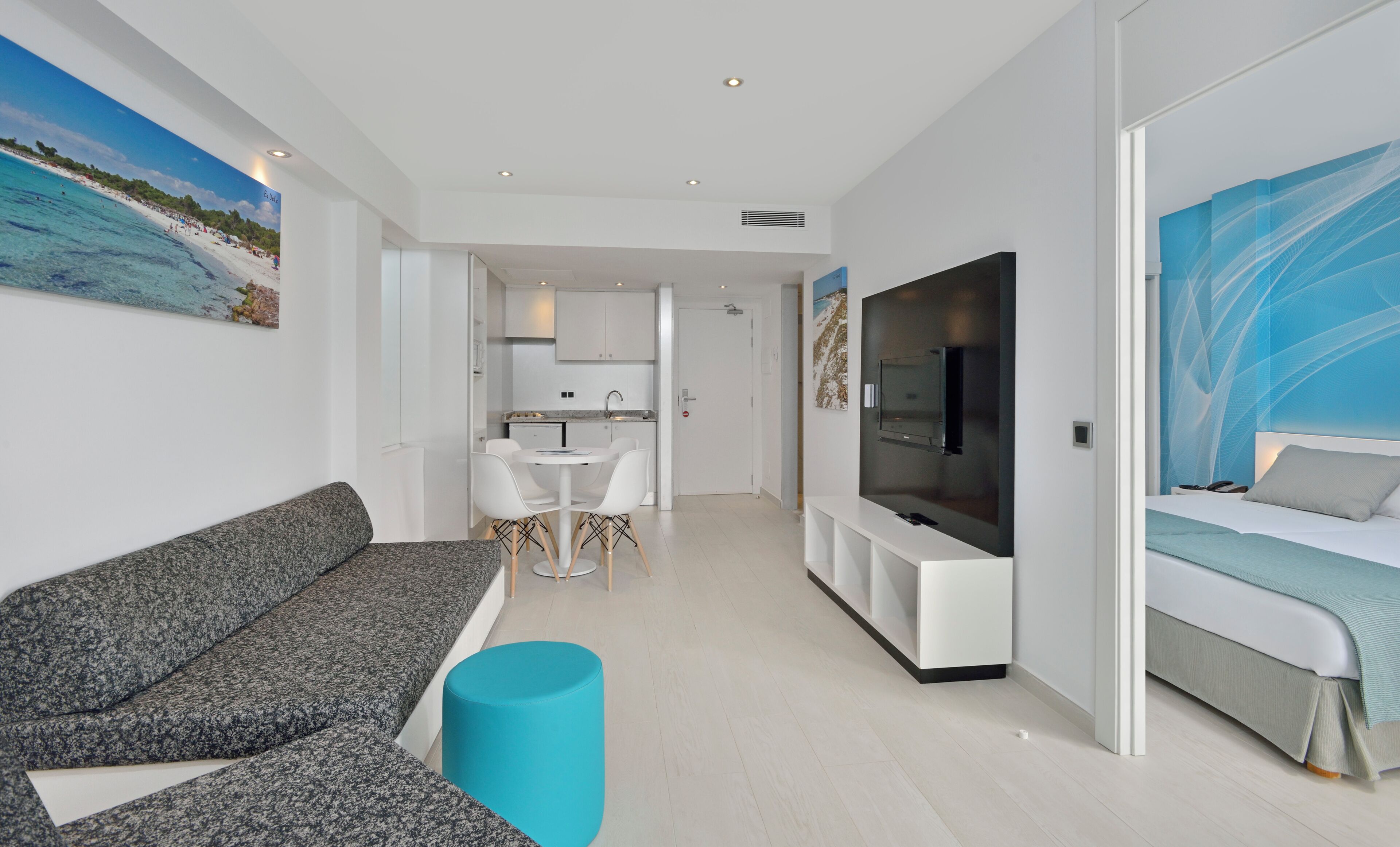 Una sala moderna con un sofá gris, un reposapiés azul, un televisor y una cama con colcha azul claro.