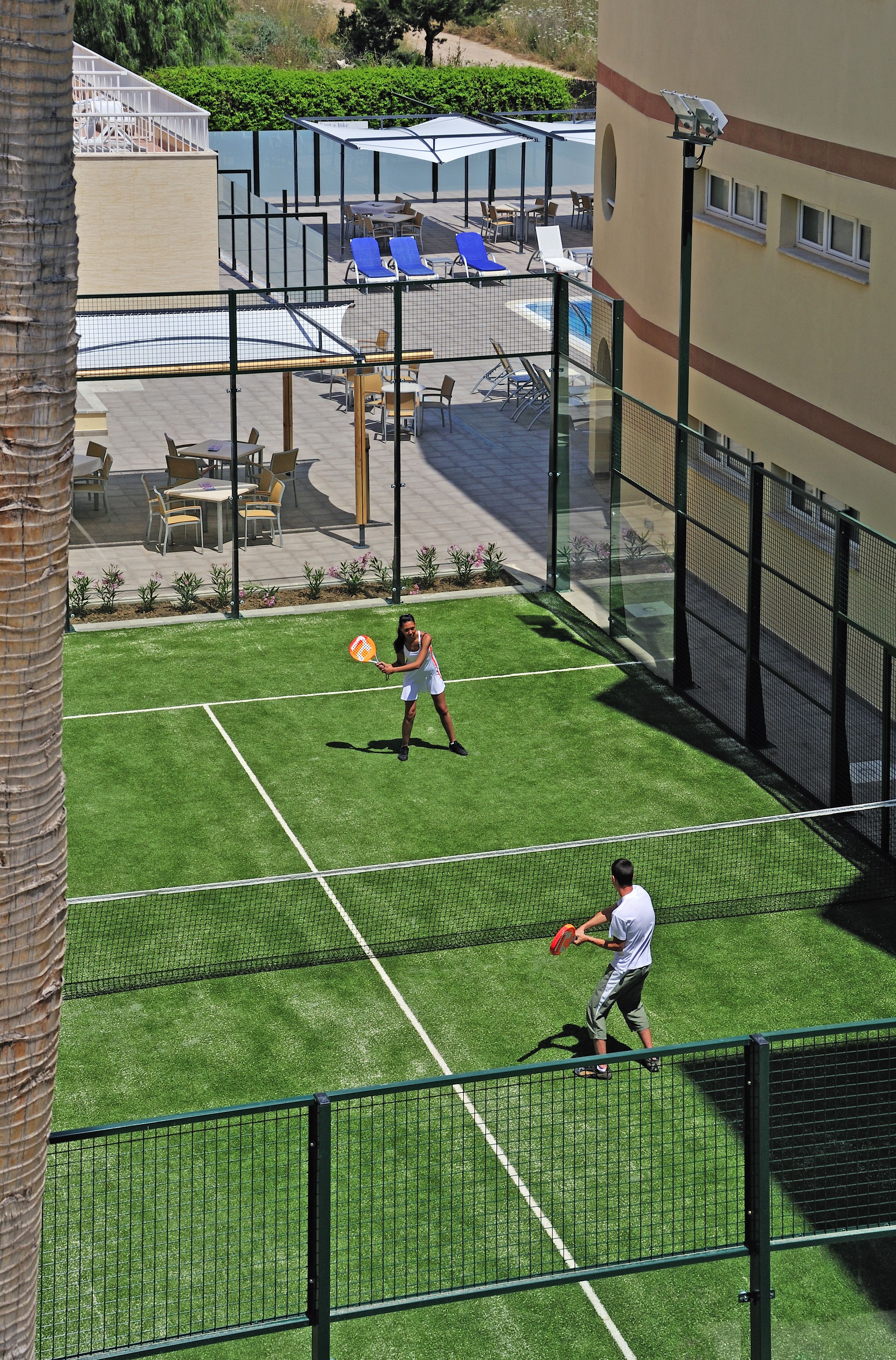 Pista de tenis al aire libre con red, rodeada por una valla y un área de piscina con tumbonas y sombrillas.