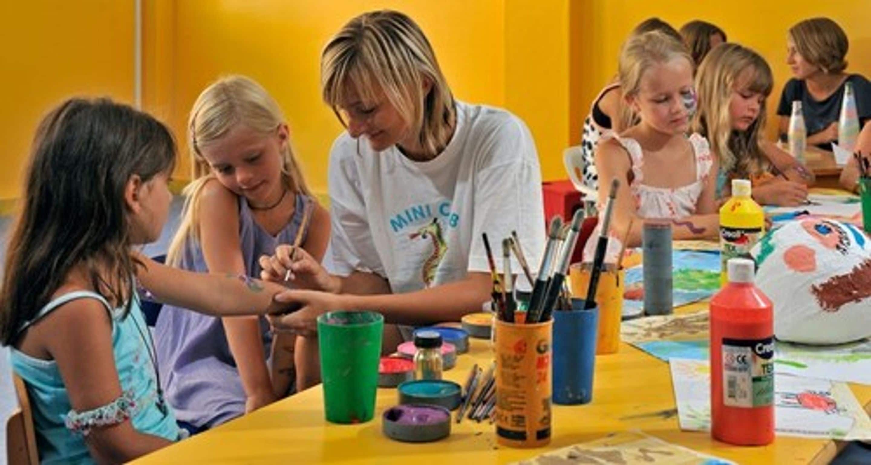 A group of children engaged in a painting activity with adult supervision, various art supplies on a table, and a yellow wall in the background.
