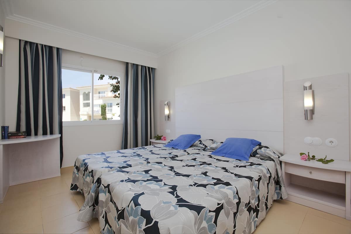 Ein Schlafzimmer mit einem großen Bett, einem Nachttisch, einem Fenster mit Vorhängen und Blick auf Gebäude.