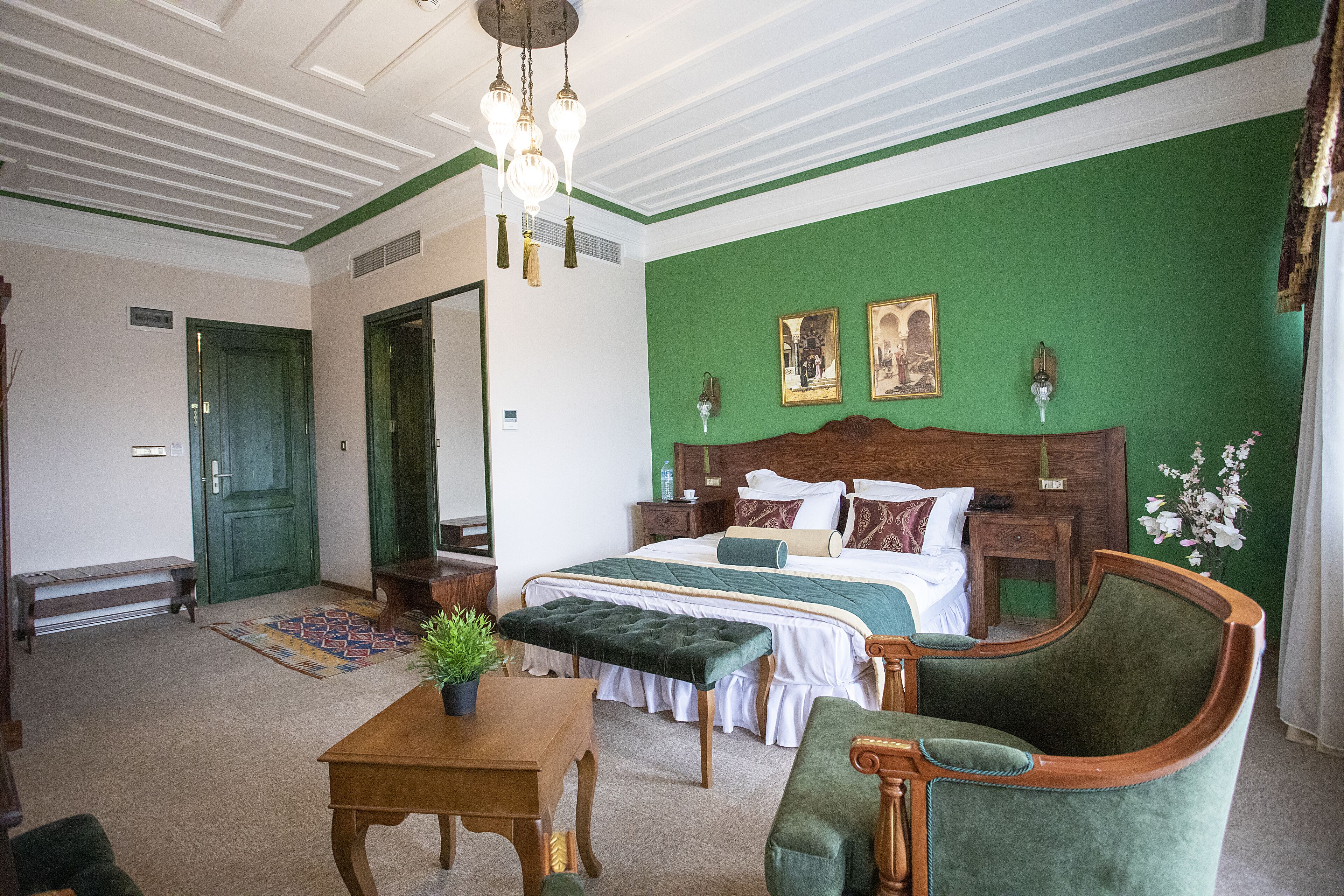 Ein Hotelzimmer mit einer grünen Akzentwand, einem großen Bett mit Holz-Kopfschild, einem grünen Polsterarmlehnstuhl, einer Holzbank und einem kleinen Tisch mit Pflanze.
