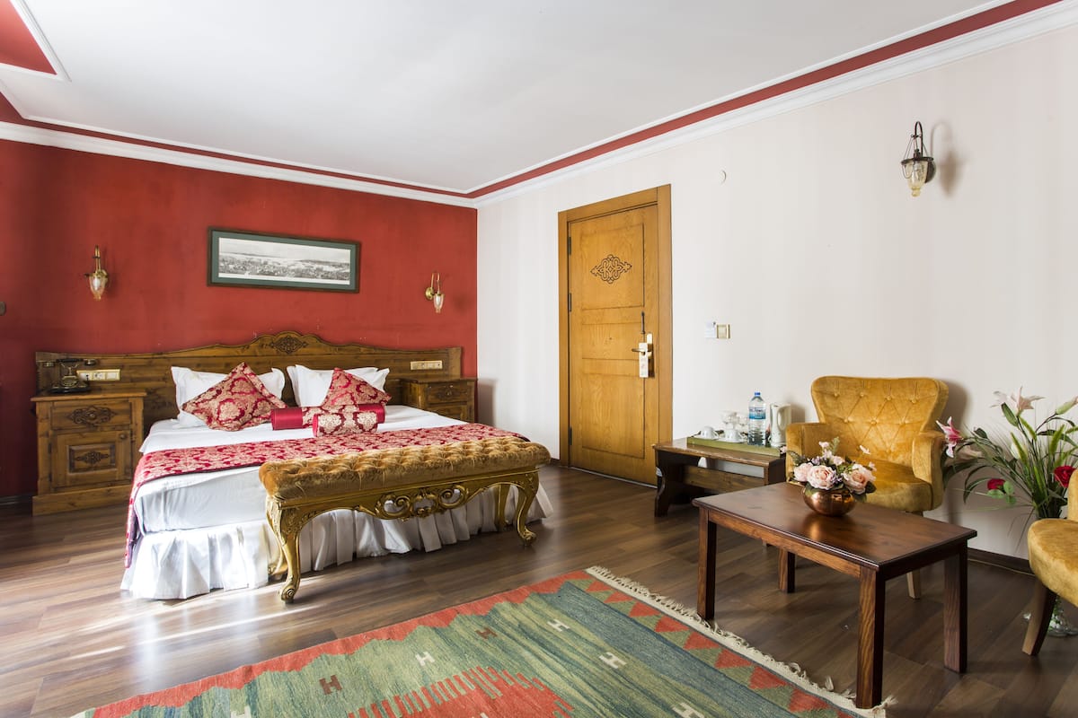 A bedroom with a wooden bed, a red accent wall, a wooden door, a small table with a vase, and a chair.