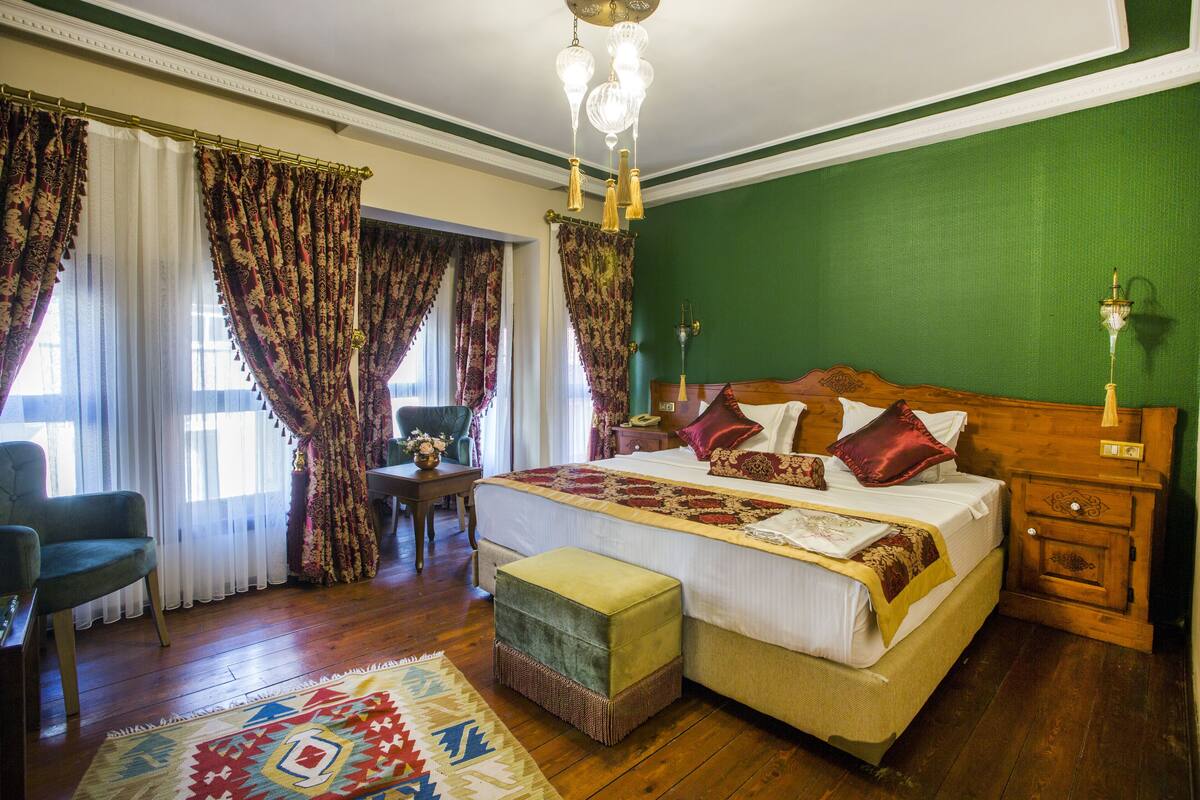 A bedroom with a large bed, a wooden nightstand, a green wall, and a patterned rug.