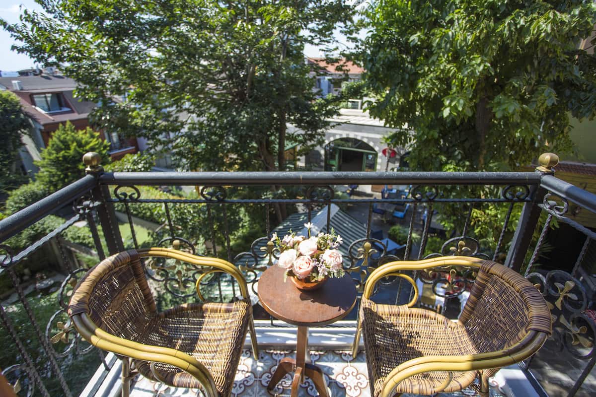 A balcony with wicker chairs and a small table, overlooking a garden and buildings.