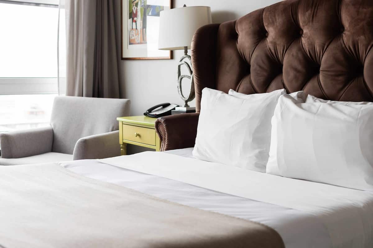 Una cama bien tendida con sábanas blancas, un cabecero marrón acolchado y dos almohadas blancas. Se ve una mesita de noche verde con una lámpara y un teléfono a la izquierda.