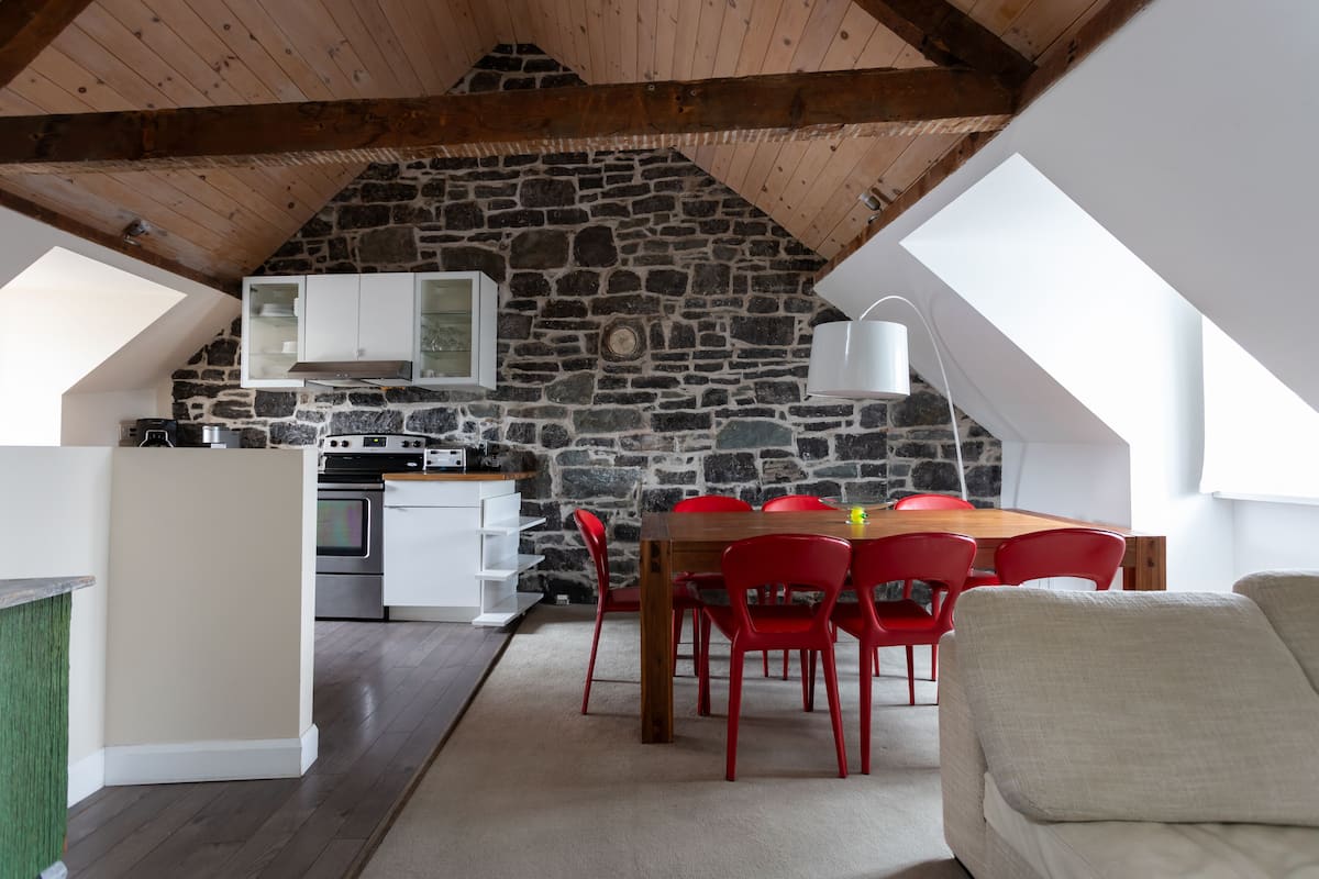 Un cuarto acogedor estilo ático con pared de piedra, vigas de madera, comedor con sillas rojas y kitchenette.