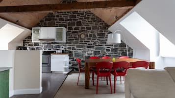 Un cuarto acogedor estilo ático con pared de piedra, vigas de madera, comedor con sillas rojas y kitchenette.